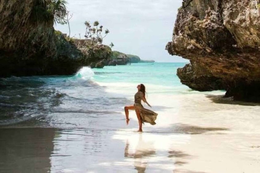 A woman standing on a beach with waves crashing onto the shore, surrounded by rocky cliffs and palm trees. 2