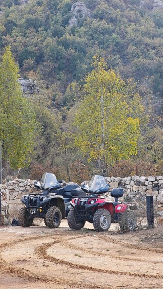 Two ATVs parked on a dirt path with a stone wall and trees in the background. 2