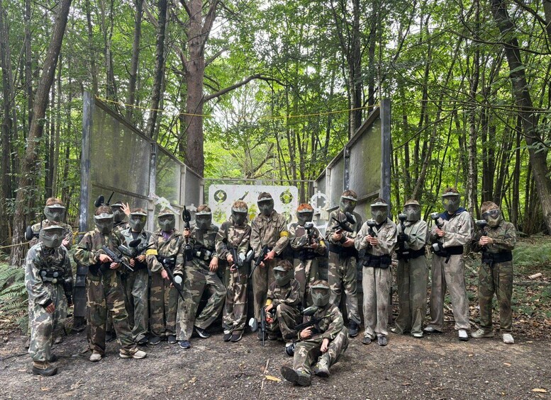A group of people in camouflage paintball gear standing in a forested area with trees and a wooden structure in the background. 4