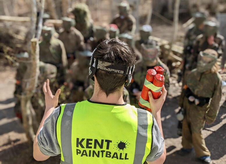 A person in a high-visibility vest holding paintball equipment, with other individuals in the background. 1