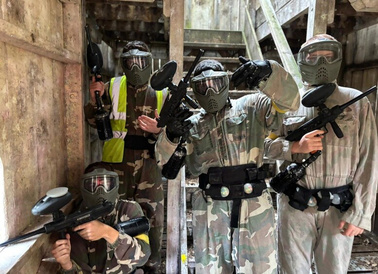 People in camouflage paintball gear with paintball guns, standing in an indoor setting with wooden structures. 5