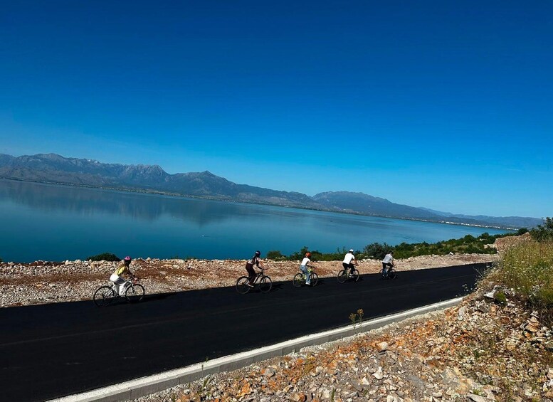 A group of cyclists on a paved road beside a calm lake with mountains in the background. 3