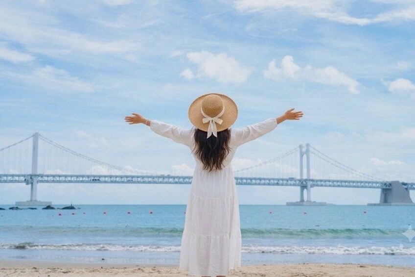A person standing on a beach with arms outstretched, facing a large bridge in the background. 1