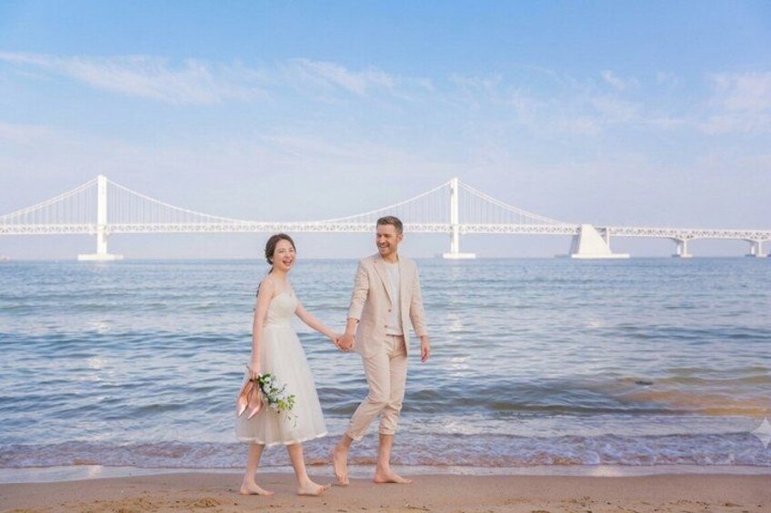 A couple holding hands on a beach with a bridge in the background. 4