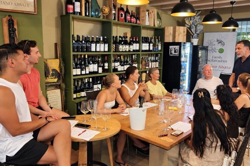 A group of people seated at a wooden table in a wine tasting room with wine bottles on shelves. 4