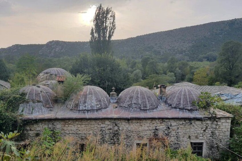 A stone building with multiple domed structures on its roof, surrounded by greenery and hills in the background. 3