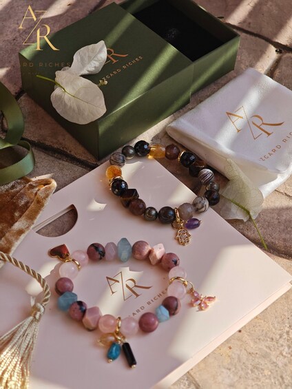 Trois bracelets ornés de différents types de perles et de breloques, une boîte verte avec une fleur blanche et un tissu blanc portant un logo. 1