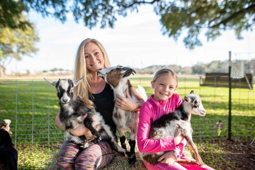 A woman and two children holding goats in a fenced outdoor area with trees and a clear sky. 3