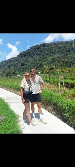 Deux personnes se tiennent sur un chemin de béton, avec un paysage verdoyant luxuriant et une colline en arrière-plan. 2