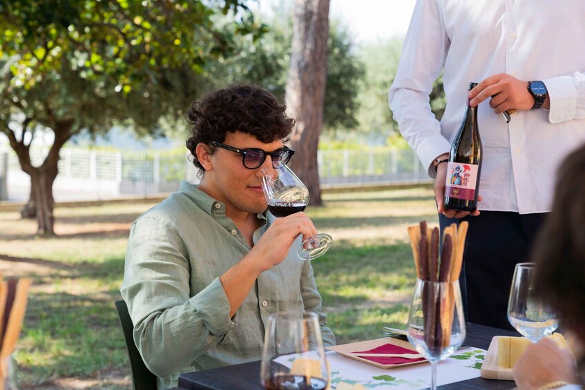 A person seated outdoors, holding a wine glass, with another person pouring wine from a bottle. 2