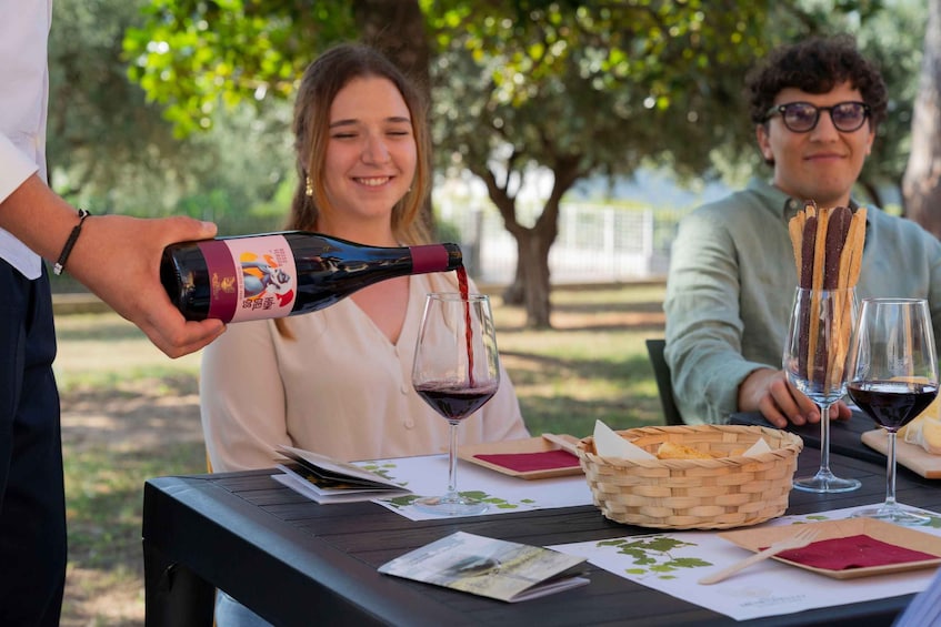 A person pouring wine into a glass, a basket of breadsticks, and a table with menus and a placemat. 1