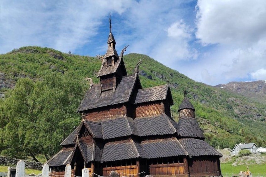 A traditional wooden church with a steeply pitched roof and multiple spires, set against a backdrop of green hills and a clear sky. 2