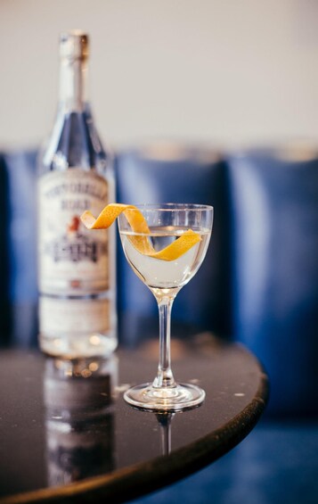 A cocktail glass with a lemon peel garnish on a dark table, a bottle of liquor, and blue upholstered seating in the background. 2