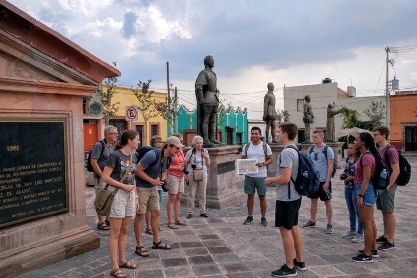 A group of people standing in a plaza with a statue and a plaque in the background. 1