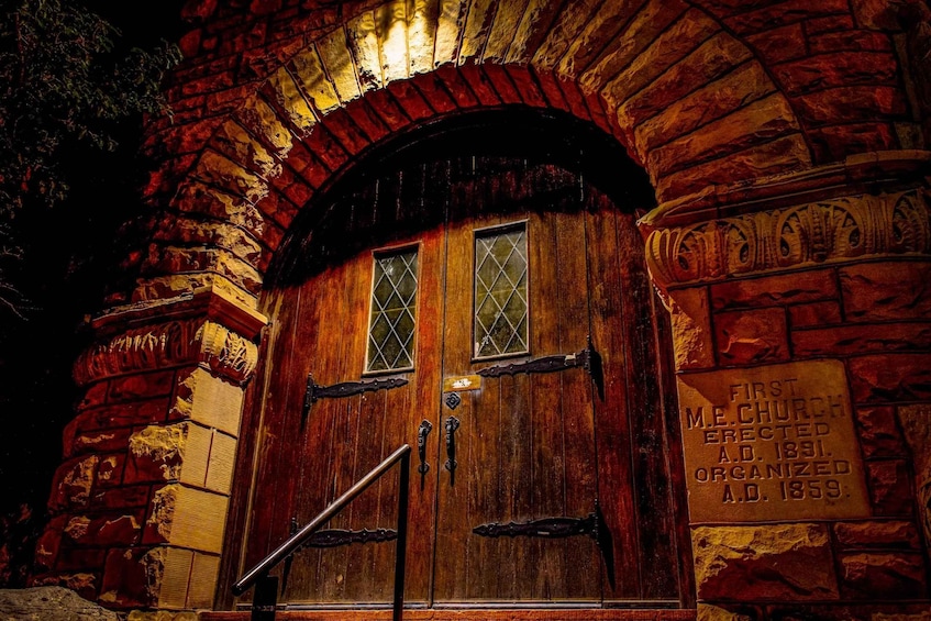 A wooden door with a metal handle and glass panes, set in a stone building with a brick archway. A sign on the wall reads 'FIRST M.E. CHURCH ERECTED A.D. 1831. ORGANIZED A.D. 1859.' 3