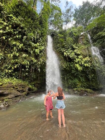 Zwei Personen stehen im flachen Wasser nahe einem Wasserfall, umgeben von dichtem Grün. 4
