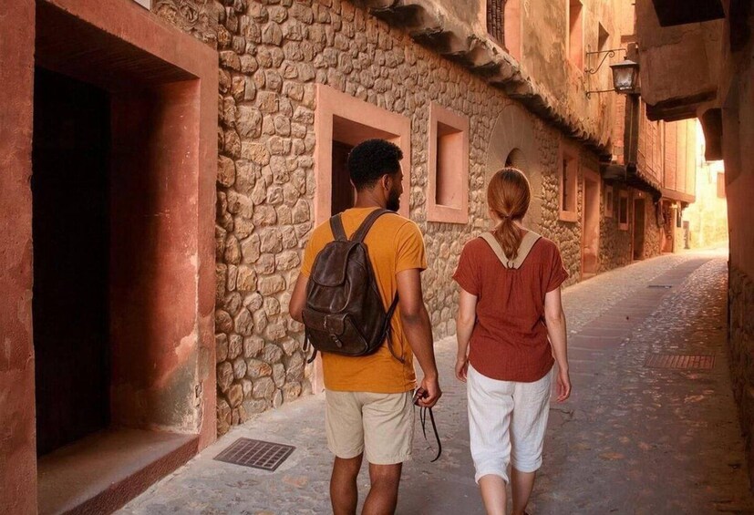 Two people walking down a narrow cobblestone street with stone walls and wooden doors. 3