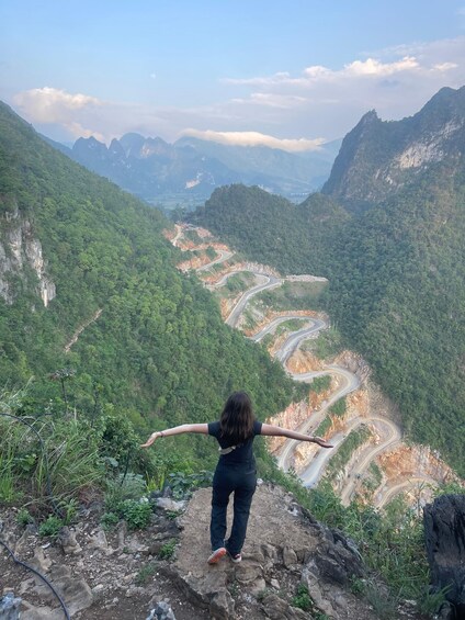 Une personne se tient debout sur un promontoire rocheux, les bras tendus, et contemple une route sinueuse qui serpente à travers un paysage montagneux. 3