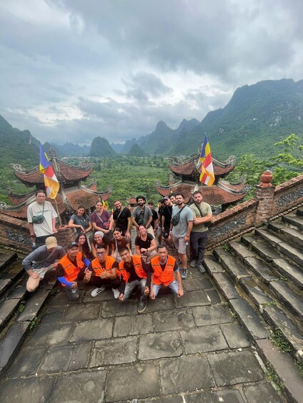 Eine Gruppe von Menschen posiert auf einer traditionellen Architekturstruktur mit Flaggen vor einer gebirgigen Landschaft. 4