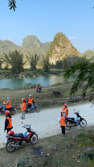 Eine Gruppe von Personen in orangefarbenen Westen versammelt sich in der Nähe eines Flusses, mit abgestellten Motorrädern und Bergen im Hintergrund. 3