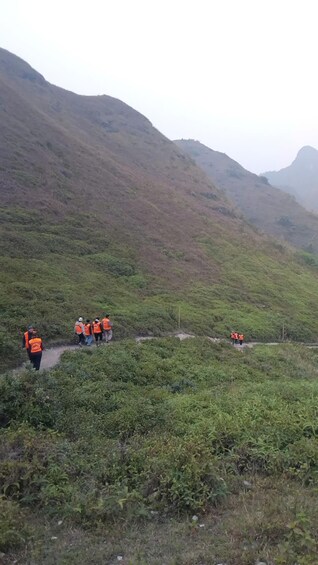 Eine Gruppe von Personen in orangefarbenen Westen geht auf einem schmalen Pfad durch eine gebirgige Landschaft mit dichter Vegetation. 5