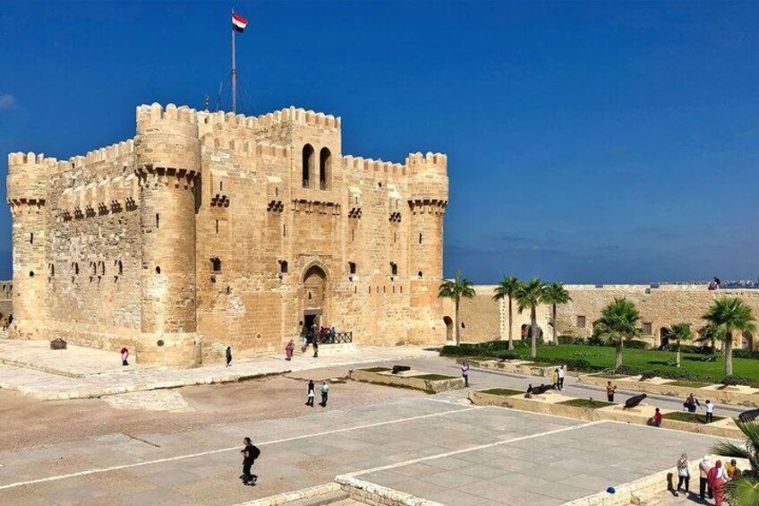 A historic stone castle with a flag, palm trees, and a paved area with people. 1