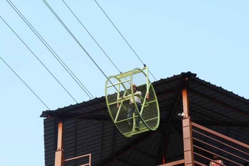 A person in a green basket suspended by cables above a building with a metal roof. 2