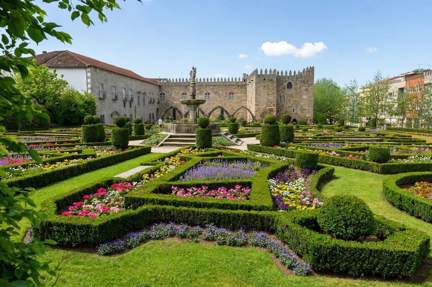 A formal garden with geometrically arranged flower beds, a central fountain, and a historic building with a crenellated tower. 3