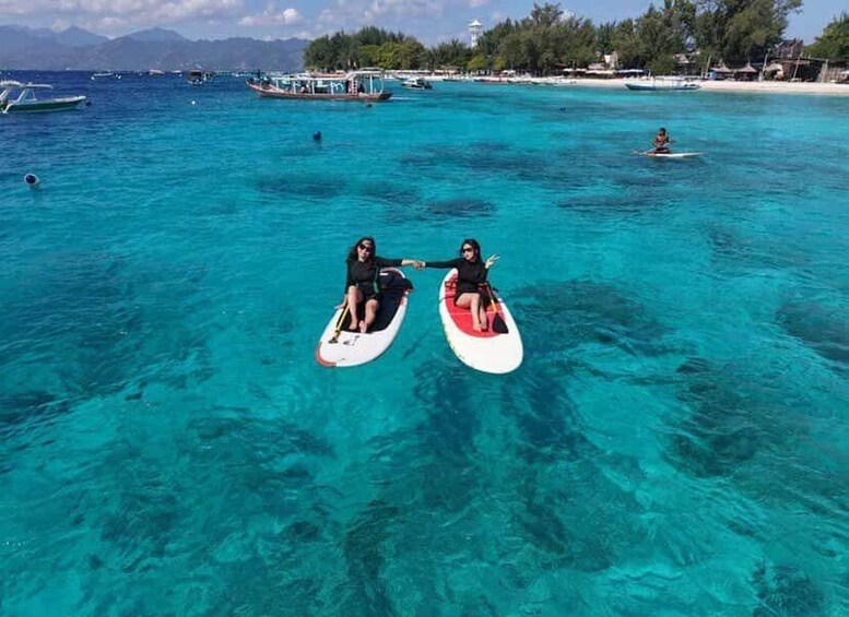 Zwei Personen auf Paddleboards in klarem türkisblauem Wasser, im Hintergrund Boote und ein Strand. 3