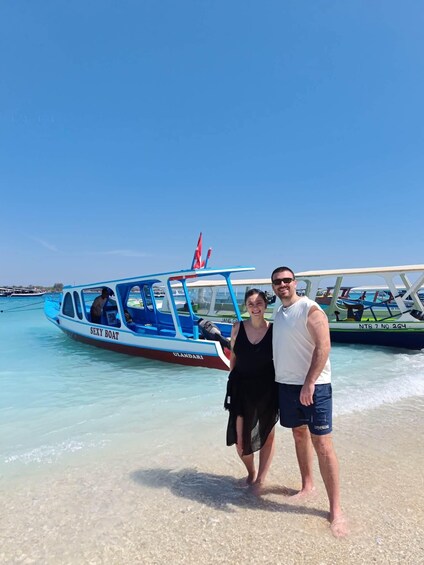 Ein Paar steht an einem Strand, im Wasser liegt ein blau-weißes Boot mit dem Namen 'SEXY BOAT'. 1