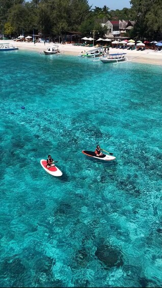 Zwei Personen paddeln auf Surfbrettern in klarem türkisblauem Wasser, im Hintergrund ein Strand und Boote. 2