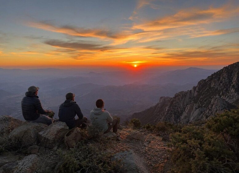 Three people sitting on a rocky outcrop overlooking a mountainous landscape during sunset. 3