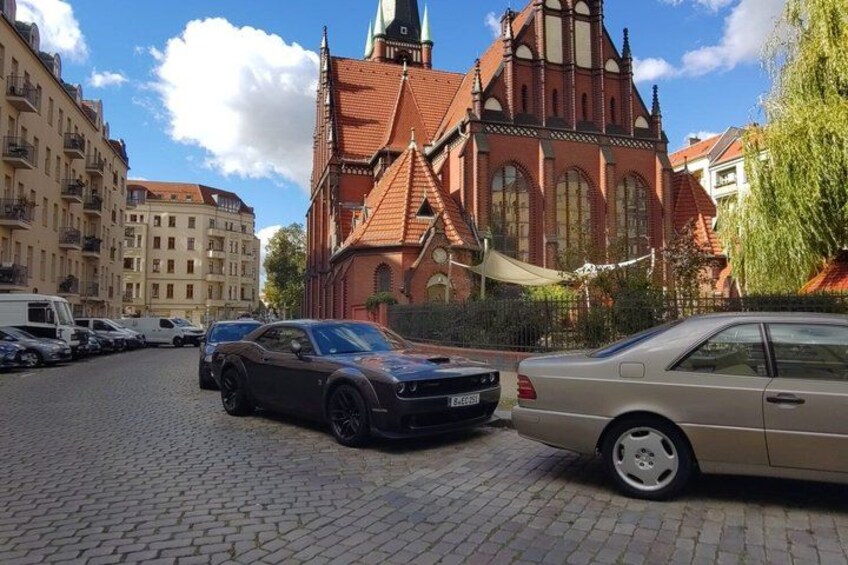 A cobblestone street, a church with a red roof, and parked cars. 2