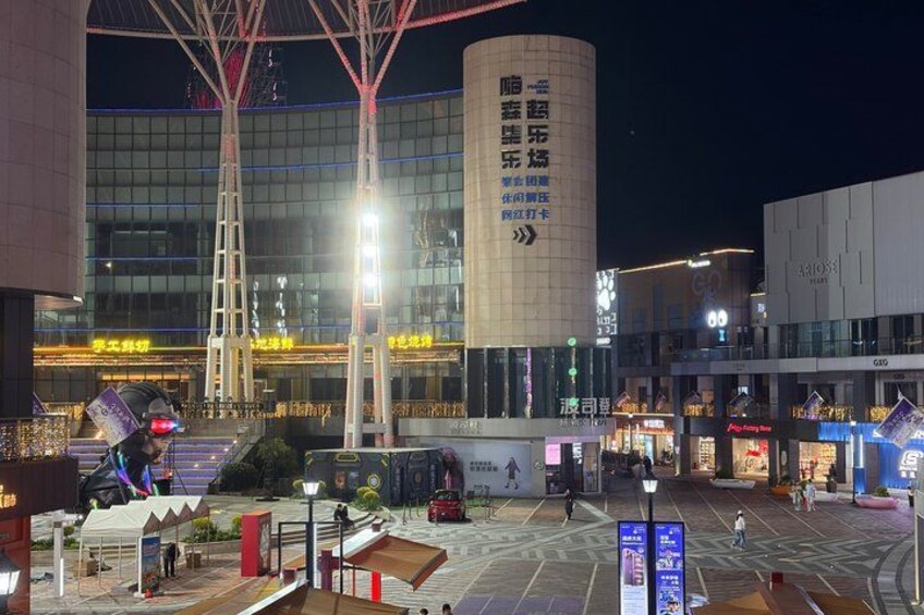 A nighttime urban square with illuminated buildings, a large cylindrical structure, and a pedestrian area with shops. 2
