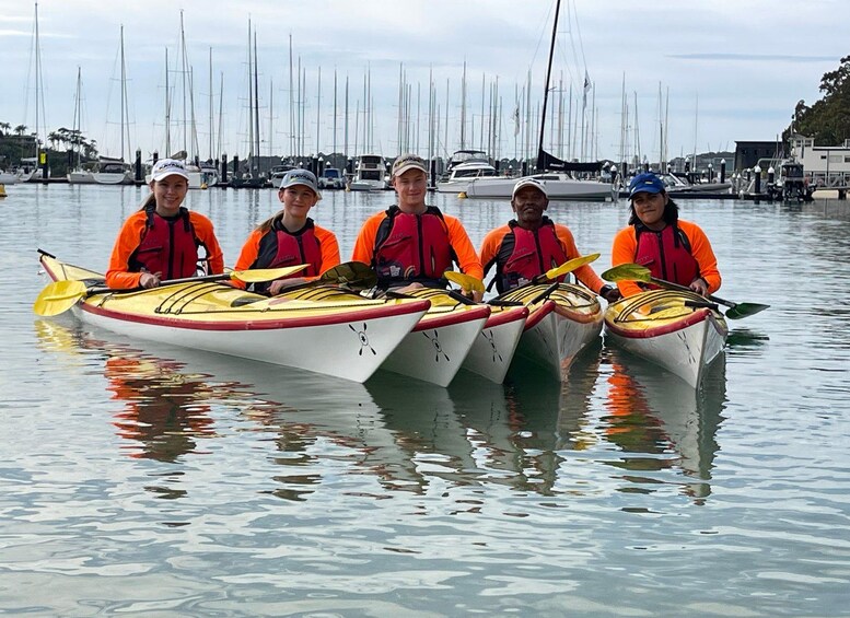 Cinq personnes en kayaks, portant des gilets de sauvetage orange, sont alignées sur une étendue d’eau calme, avec des voiliers en arrière-plan. 2