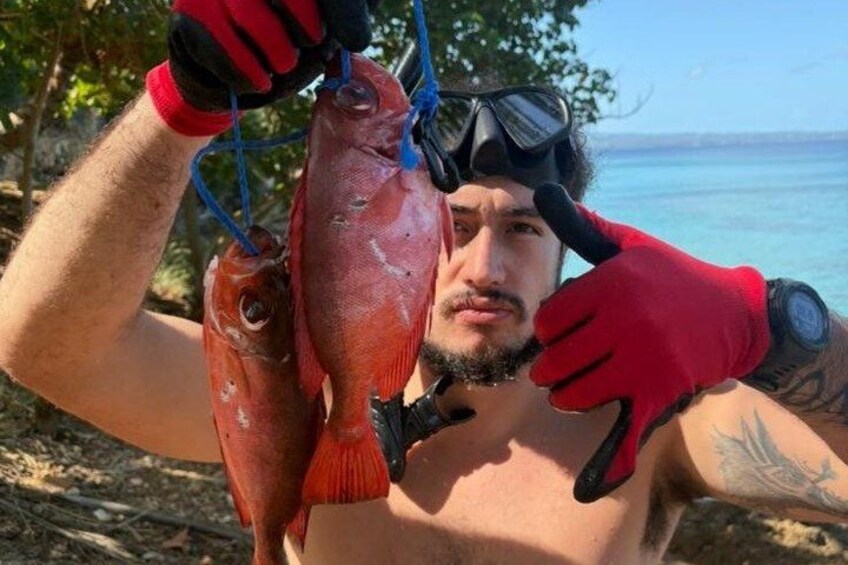 A person holding two fish, wearing red gloves and a watch, with a clear sky and ocean in the background. 1