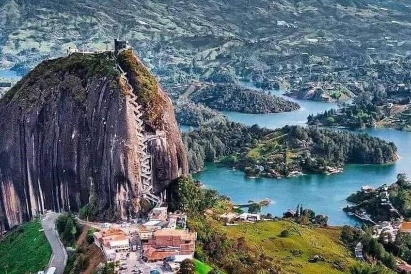 A large rock formation with a staircase leading up to a peak, surrounded by a lake and a village. 4