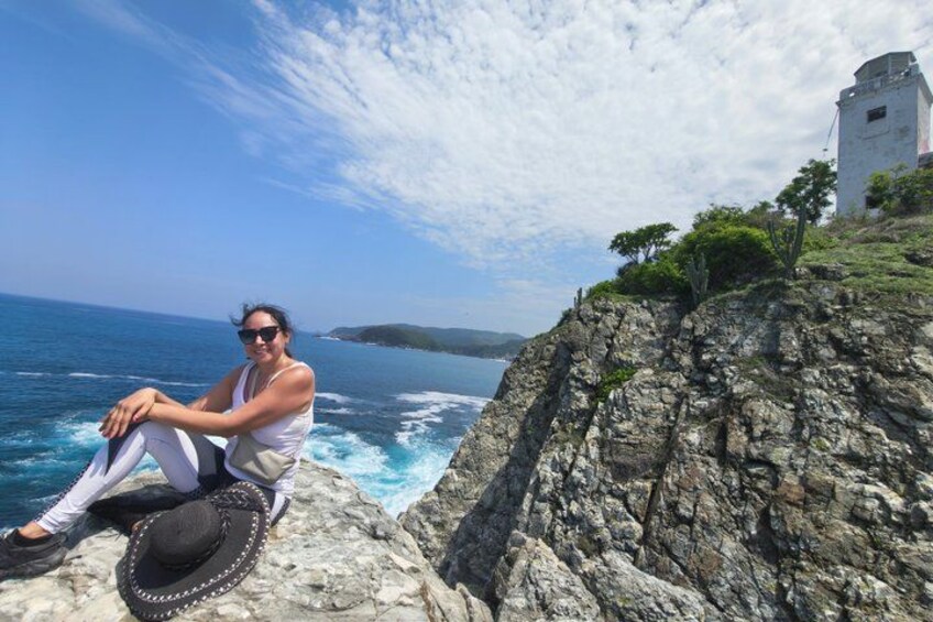 A person sitting on a rocky outcrop by the sea, with a lighthouse in the background. 2