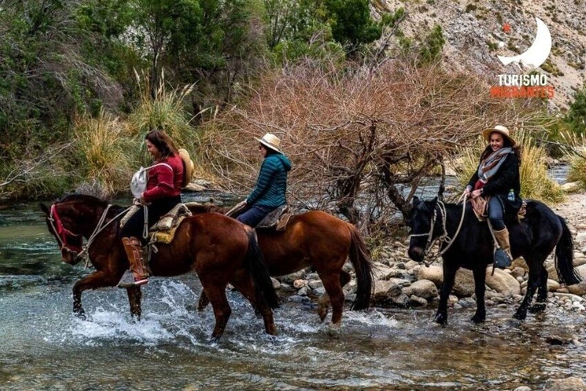 Three people riding horses across a shallow river, surrounded by rocky terrain and sparse vegetation. 3