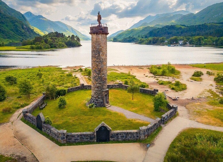 Une tour de pierre surmontée d’une statue, entourée d’un mur circulaire en pierre et d’une zone herbeuse, avec des montagnes en arrière-plan. 2
