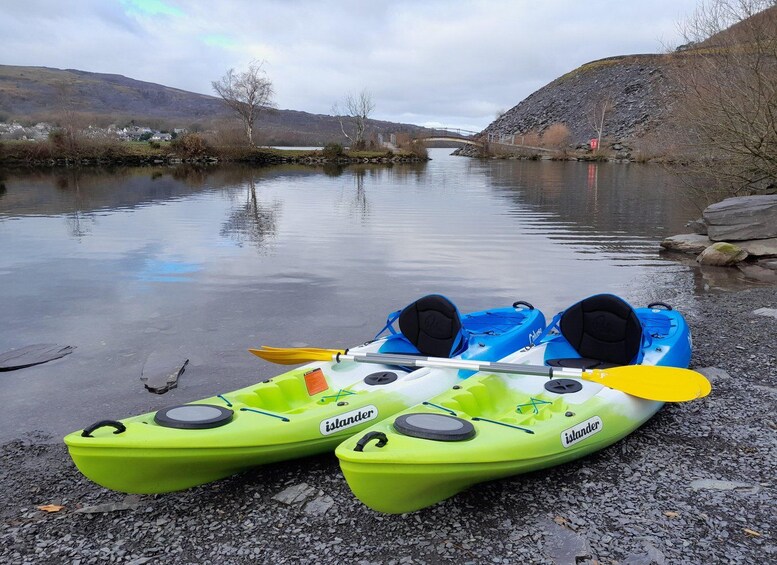 Two kayaks, one green and one blue, on a gravel shore by a calm river with a bridge in the background. 1