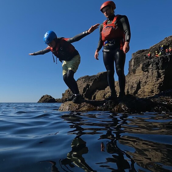 Two people in water safety gear preparing to jump into the water from a rocky cliff. 4