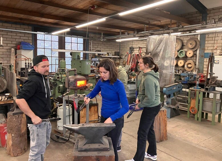 Three people in a workshop with metalworking equipment, including an anvil and a forge. 1
