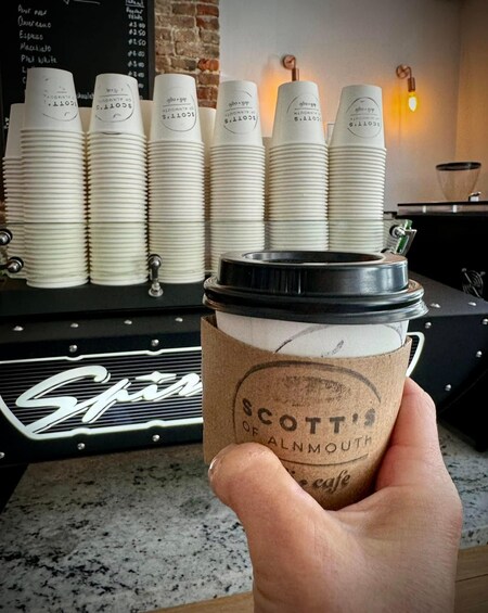 A hand holding a coffee cup with a lid, labeled 'Scott's of Alnmouth.' In the background, there are stacks of white cups and a coffee machine. 4