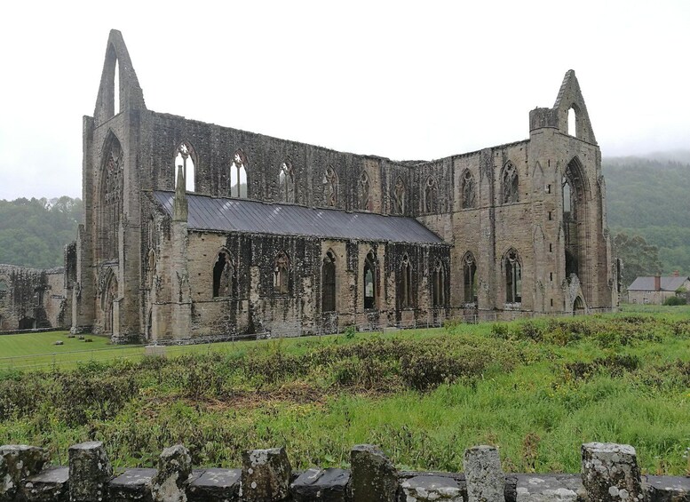An ancient stone building with arched windows and a steeply pitched roof, surrounded by a grassy field and a stone wall. 3