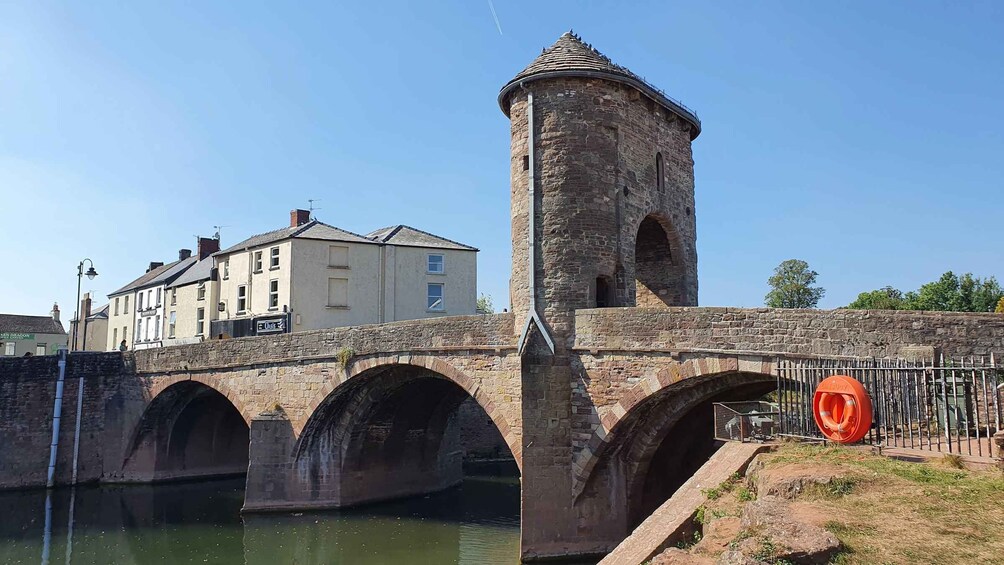 A stone bridge with arches over a body of water, a tower with a conical roof, and a building with a sign. 2
