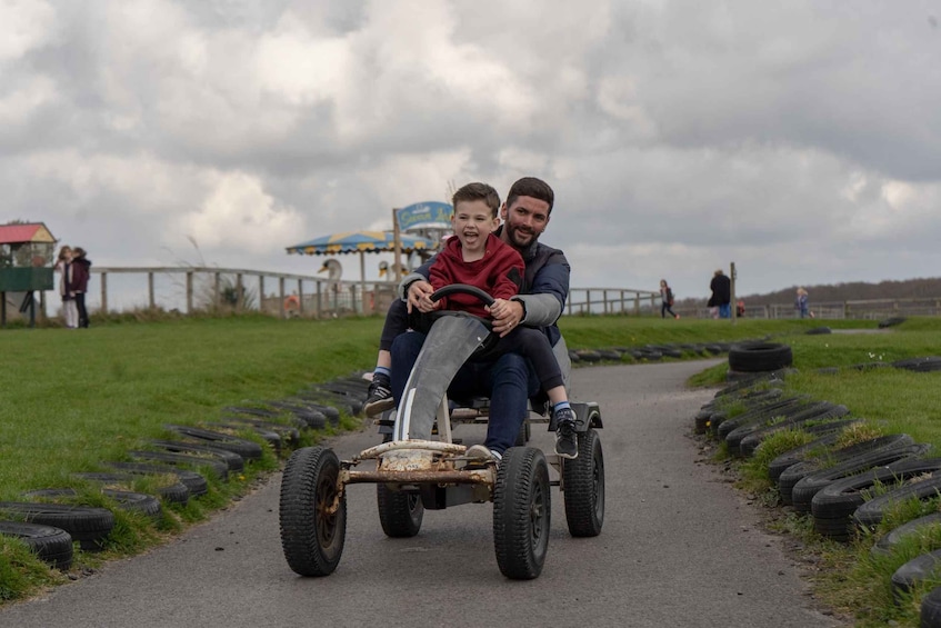 A man and a child on a rusty, makeshift vehicle with large wheels, driving on a path surrounded by grass and scattered tires. 4