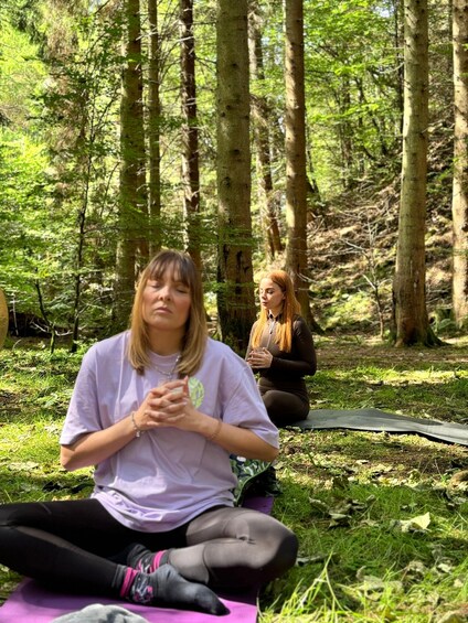 Two people sitting on mats in a forest with trees and grass. 4