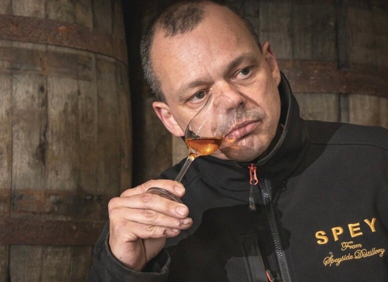 A person holding a glass of amber liquid, wearing a black jacket with 'SPEY' text. 5