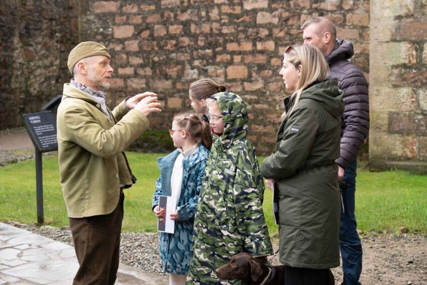 A group of people standing outdoors, engaged in a discussion, with a stone wall and a signboard in the background. 1